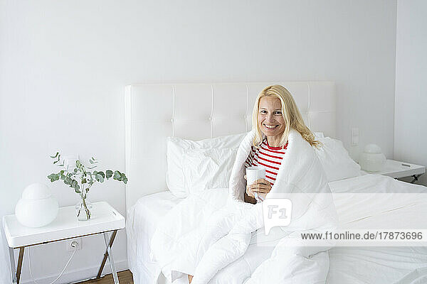 Smiling woman wrapped in blanket drinking tea in bed