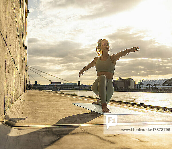 Smiling woman with arms outstretched practicing yoga on promenade at sunset