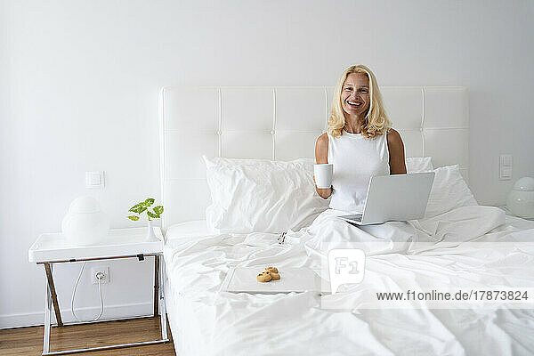 Glückliche Frau sitzt mit Laptop und Tee auf dem Bett