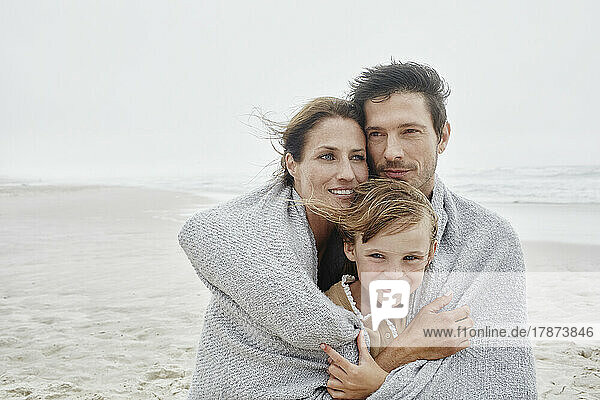 Man and woman standing with daughter on windy beach wrapped in a blanket