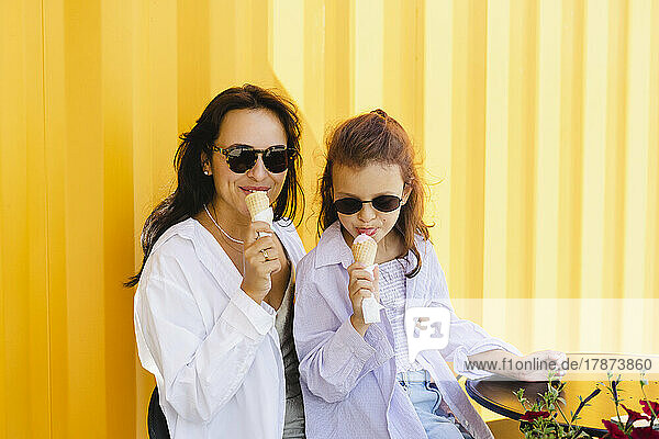 Mother and daughter wearing sunglasses eating ice cream in front of yellow wall