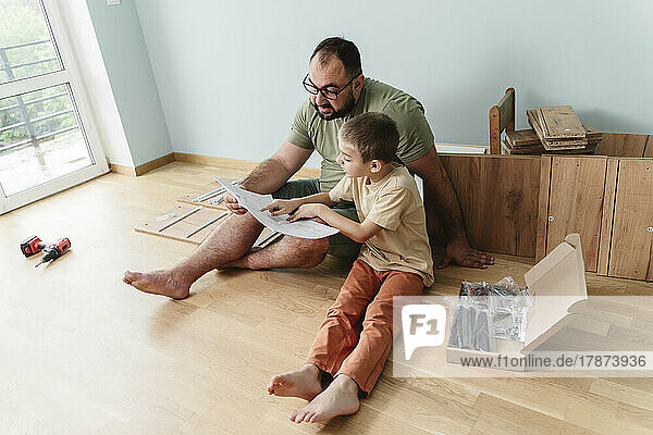 Son and father reading instruction manual sitting on floor at home