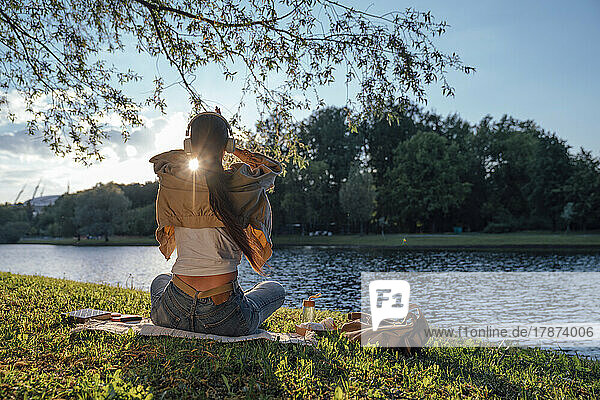 Woman sitting on grass with headphones by lake at park