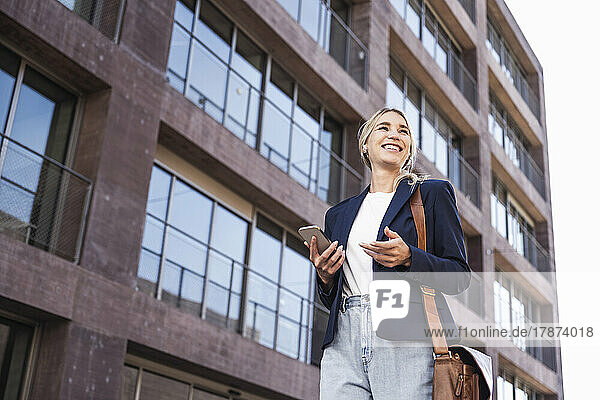Happy woman with bag and smart phone standing in front of building
