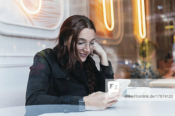 Smiling young woman using smart phone sitting at cafe