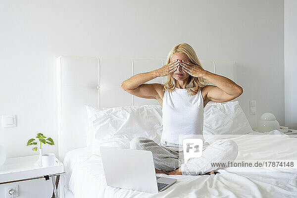 Stressed woman covering eyes sitting by laptop on bed