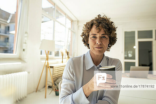 Mature businesswoman with disposable coffee cup sitting in office