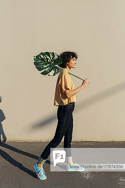 Woman holding monstera leaf standing in front of wall on sunny day