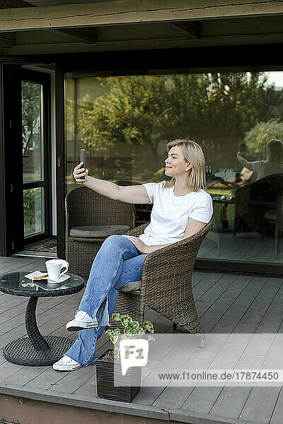 Smiling woman taking selfie through smart phone sitting on chair at porch