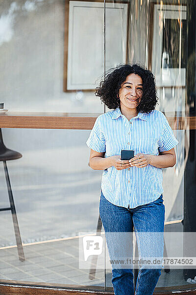 Smiling woman with smart phone leaning on glass wall
