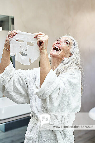 Mature woman with facial mask laughing in bathroom at home