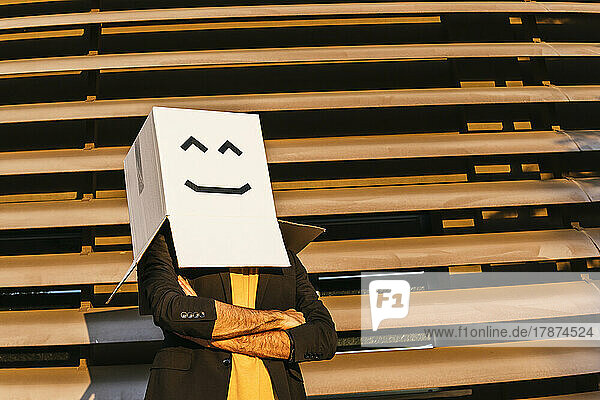 Man wearing box with smiley face standing arms crossed on sunny day