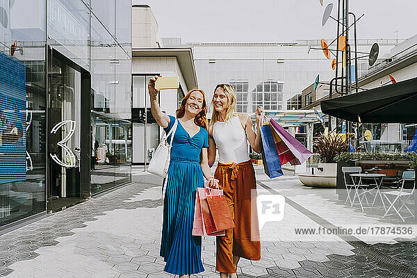 Happy friends with shopping bags taking selfie with smart phone in city