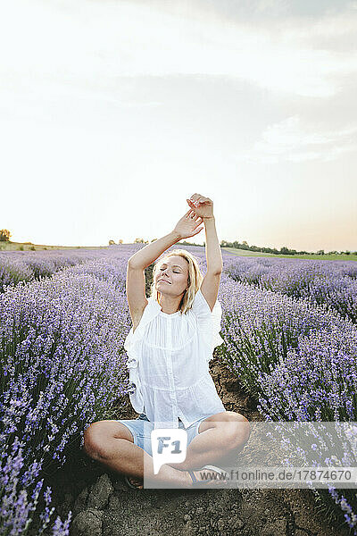 Smiling woman with arms raised sitting amidst lavender field