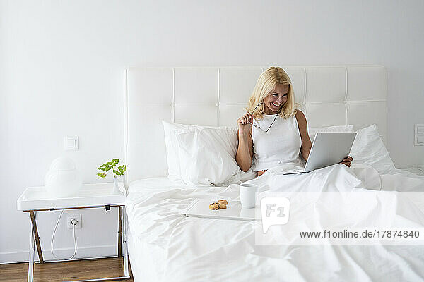 Smiling woman working on laptop by breakfast on bed