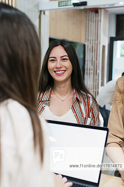 Lächelnde Geschäftsfrau schaut Kollegen mit Laptop im Büro an