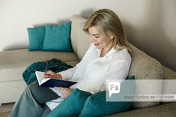 Smiling mature woman writing in diary sitting on couch at home