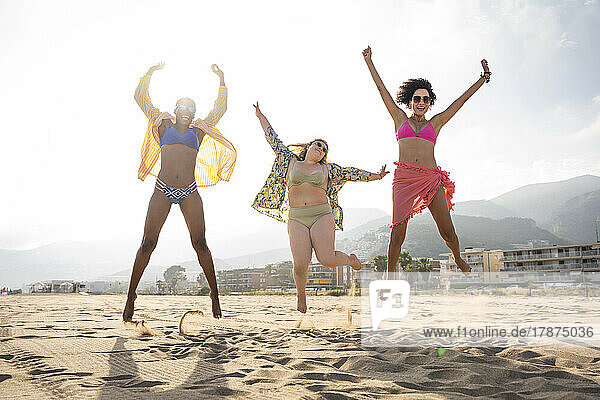 Carefree friends with arms raised jumping at beach on sunny day