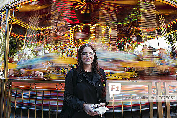 Smiling young woman standing in front of carousel at amusement park