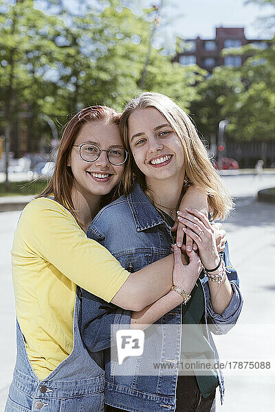 Smiling young woman embracing friend from behind on footpath