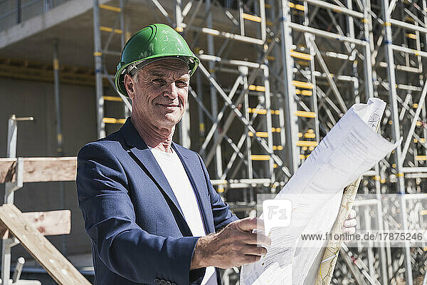 Smiling architect with blueprint standing at construction site