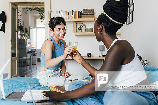 Smiling woman giving juice glass to girlfriend sitting on bed at home