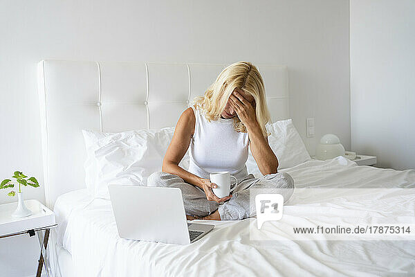 Stressed woman sitting with laptop having tea on bed