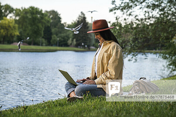 Smiling woman using laptop sitting by lake at park