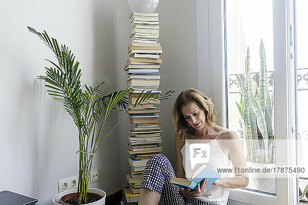 Mature woman reading book sitting by window at home