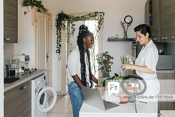 Smiling young woman talking with friend watering plant at home