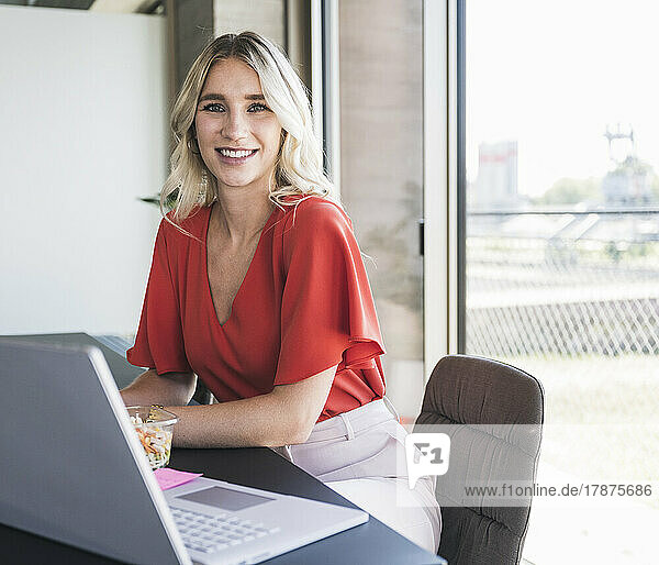 Lächelnde blonde Geschäftsfrau sitzt auf einem Stuhl im Büro