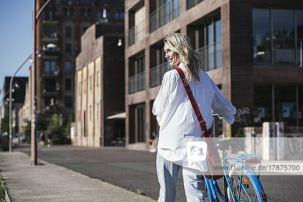 Smiling woman walking with bicycle in front of building on sunny day