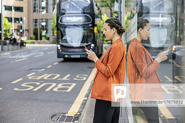 Businesswoman using smart phone leaning on glass wall at sidewalk