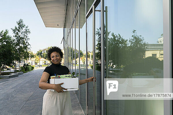 Smiling businesswoman with box standing by glass door