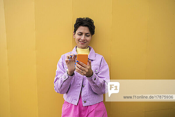 Smiling woman using smart phone in front of yellow wall