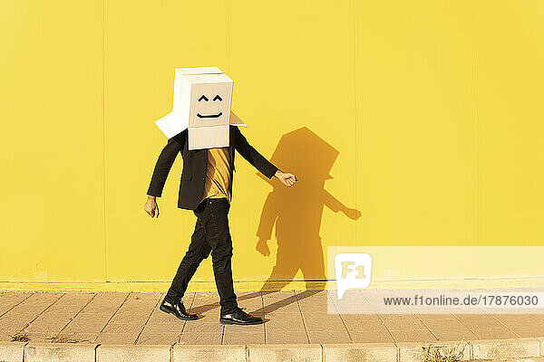 Man wearing box with face walking by yellow wall on footpath