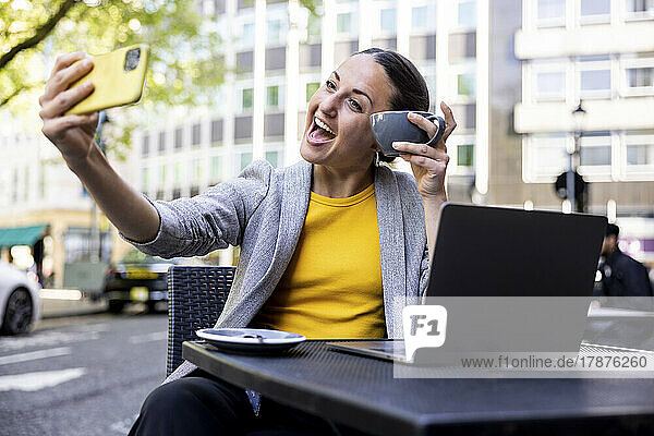 Happy businesswoman holding coffee cup taking selfie through smart phone at sidewalk cafe