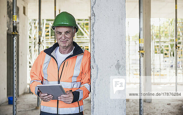 Smiling contractor with tablet PC leaning on column