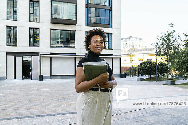 Smiling young businesswoman holding laptop in front of office building