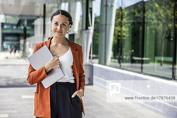 Smiling businesswoman holding laptop on footpath