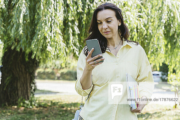 Woman with laptop and diary using smart phone in park