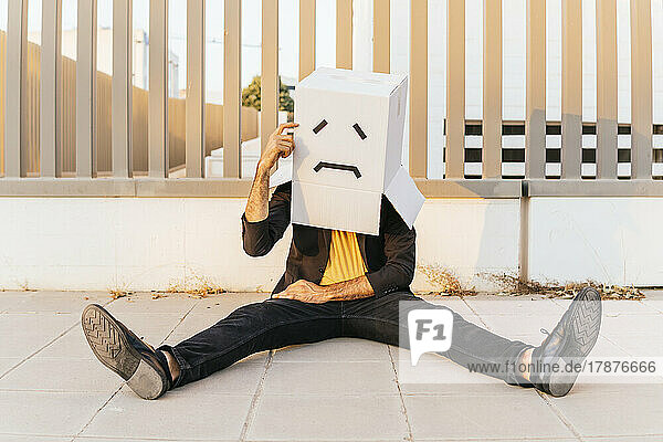 Thoughtful man wearing box with sad face sitting on footpath