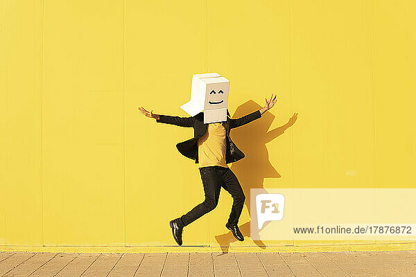 Carefree man wearing box with smiley face jumping in front of yellow wall on footpath