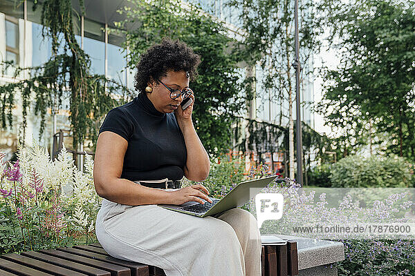 Businesswoman using laptop and talking on mobile phone sitting on bench at office park