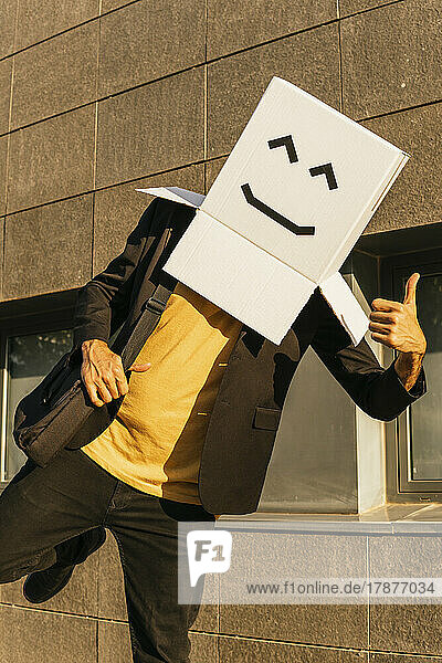Businessman wearing box with smiley face showing thumbs up on sunny day