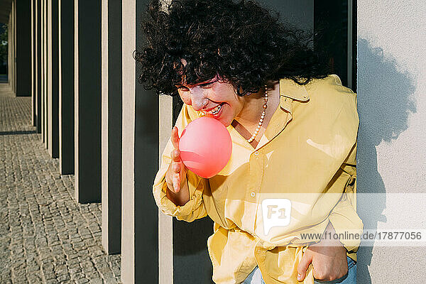 Smiling woman blowing bubble gum by column on sunny day