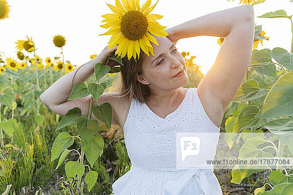 Lächelnde Frau mit erhobenen Armen und Sonnenblume auf dem Kopf