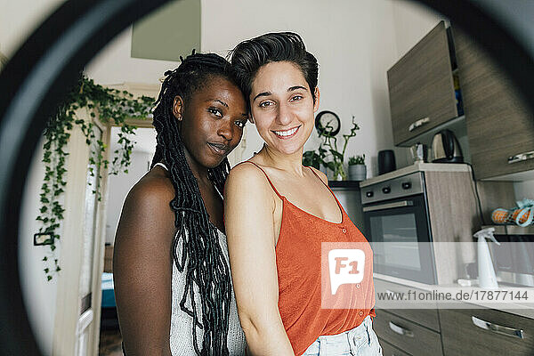 Smiling lesbian couple standing at home seen through ring light