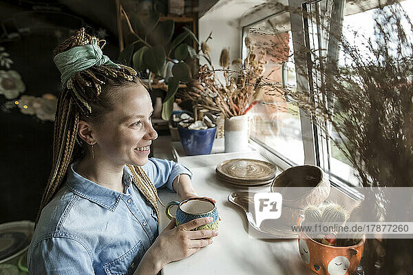 Happy potter with ceramic cup looking through window at workshop