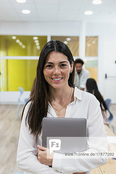 Portrait of smiling businesswoman holding laptop in office with colleagues in background